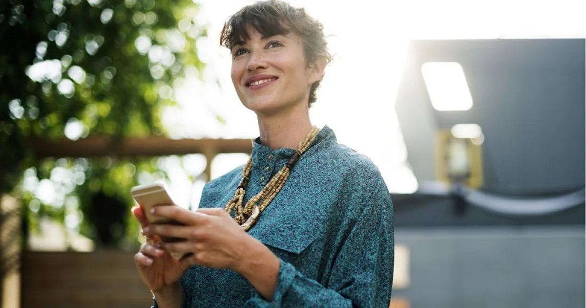 Mujer joven sonriendo mientras consulta una aplicación de bienestar en su smartphone al aire libre, representando el uso de la tecnología como aliada para mejorar la calidad de vida y la salud mental.