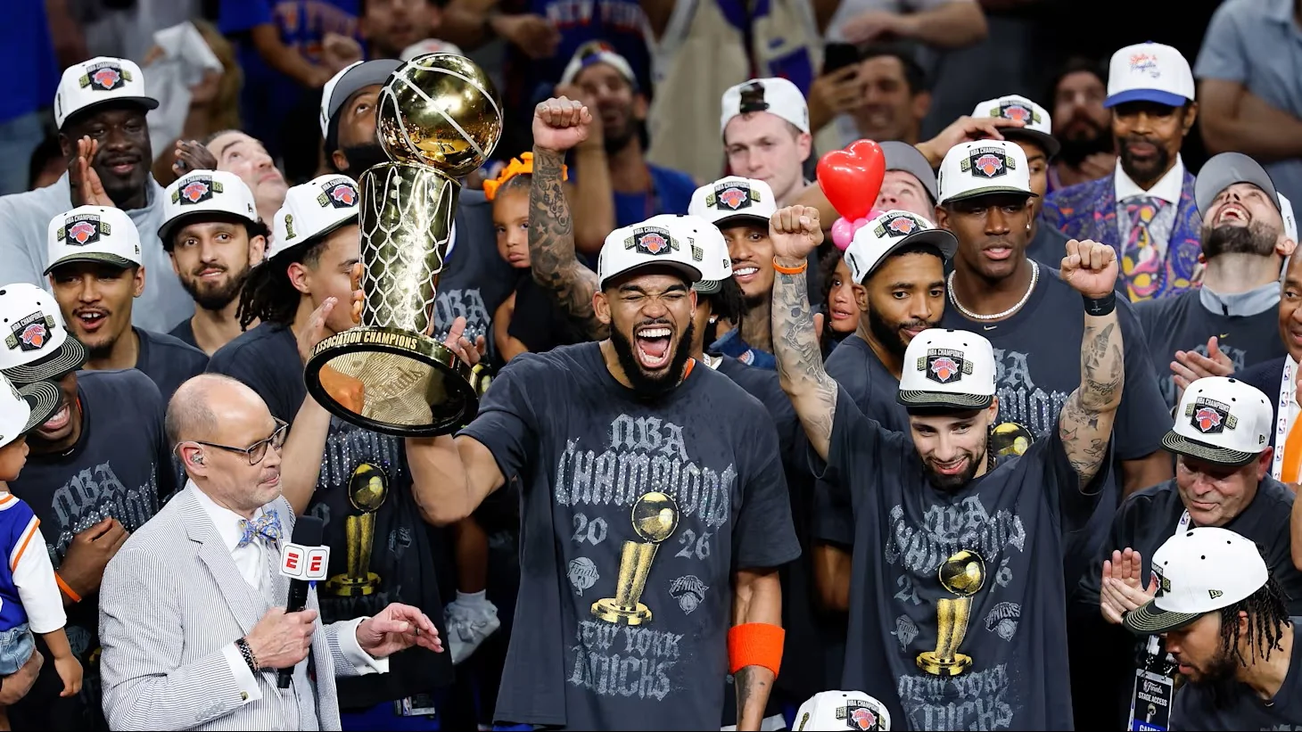 Jugadores y cuerpo técnico de los New York Knicks celebrando con entusiasmo la consecución del campeonato de la NBA 2026. En el centro, Karl-Anthony Towns sostiene el trofeo Larry O'Brien dorados con la mano derecha mientras grita eufórico con el puño izquierdo en alto. Los integrantes del equipo lucen camisetas oscuras con la inscripción «NBA CHAMPIONS 2026 NEW YORK KNICKS» y gorras blancas conmemorativas de campeones durante la entrega del título.